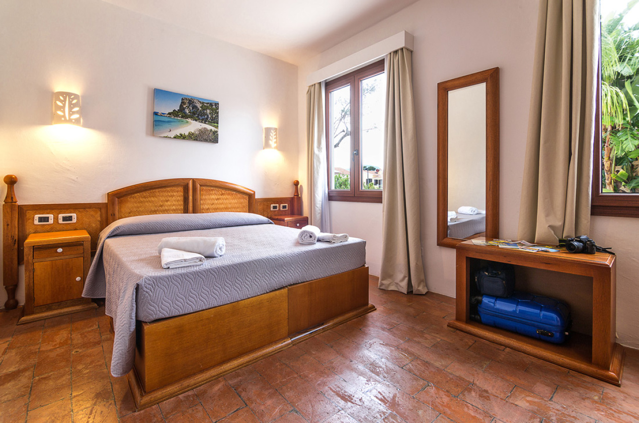Camera accogliente con letto matrimoniale, specchio, valigia e finestra luminosa.