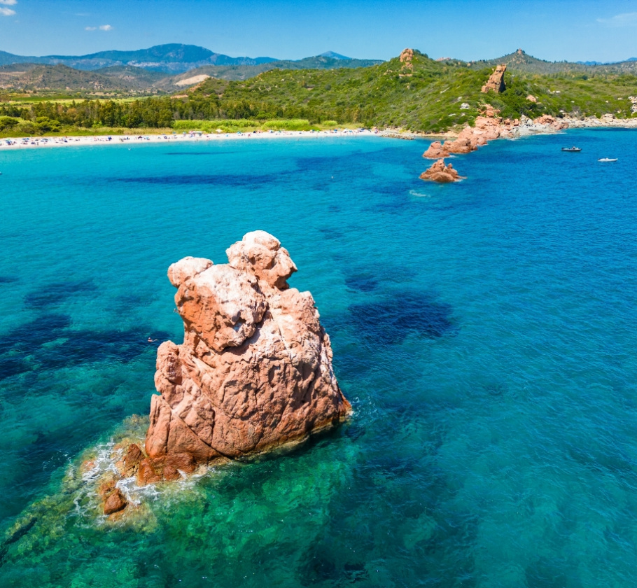Scogliera rocciosa in mare cristallino, spiaggia e colline verdi sullo sfondo.