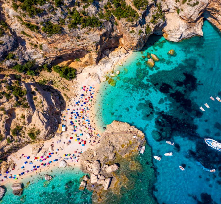 Spiaggia con ombrelloni colorati e mare cristallino, circondata da scogliere e barche.